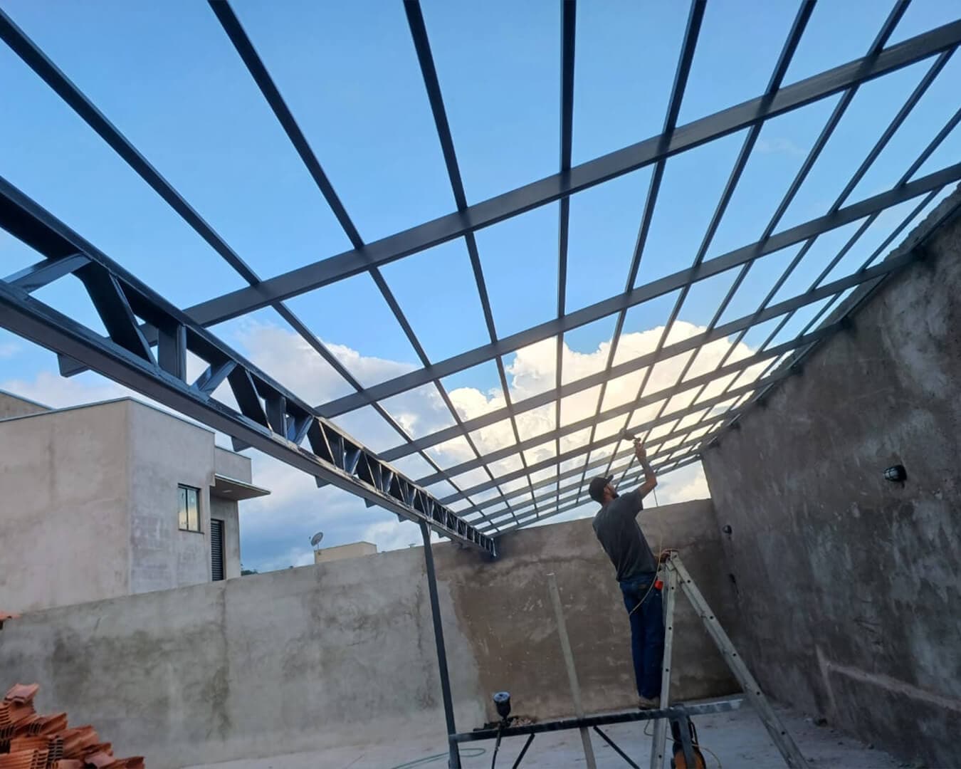 Profissional em escada soldando estrutura metálica de telhado sobre paredes de alvenaria em obra residencial